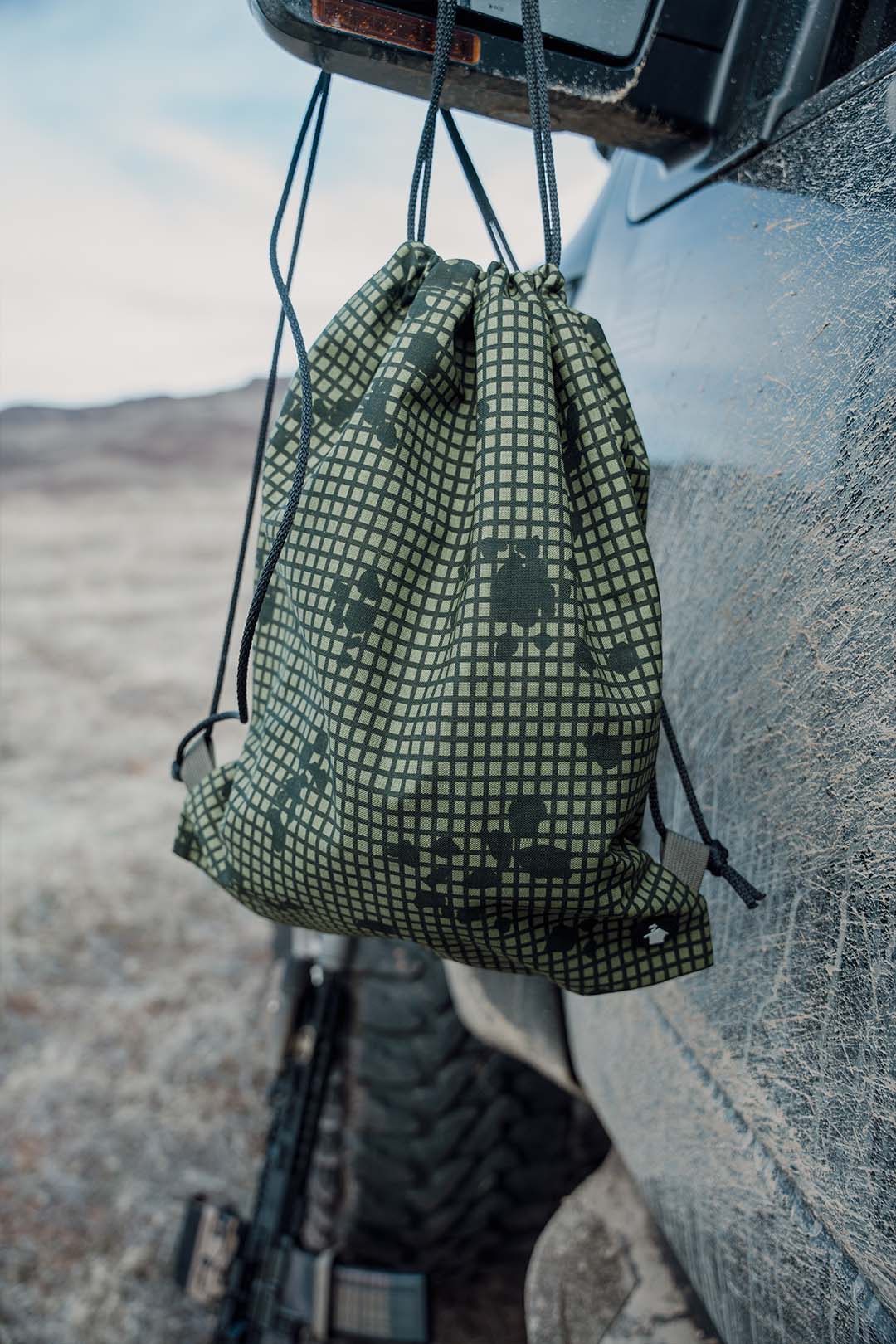 Green checkered drawstring bag hanging on a vehicle's rearview mirror with a mountainous landscape in the background.