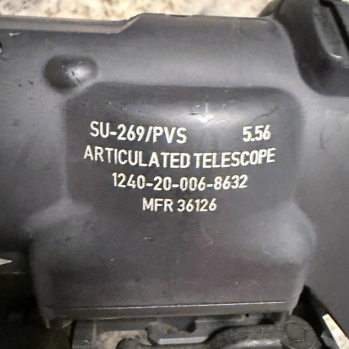 Close-up of a black articulated telescope mounting bracket with white printed part numbers and text on a speckled stone surface.