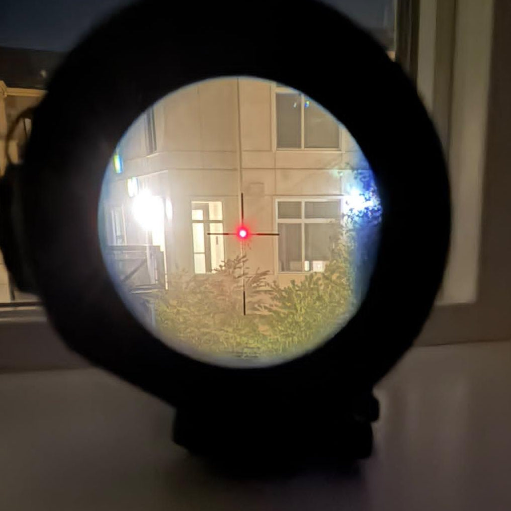 A black circular sight or scope with a red crosshair reticle, photographed on a dark surface near a window at night.