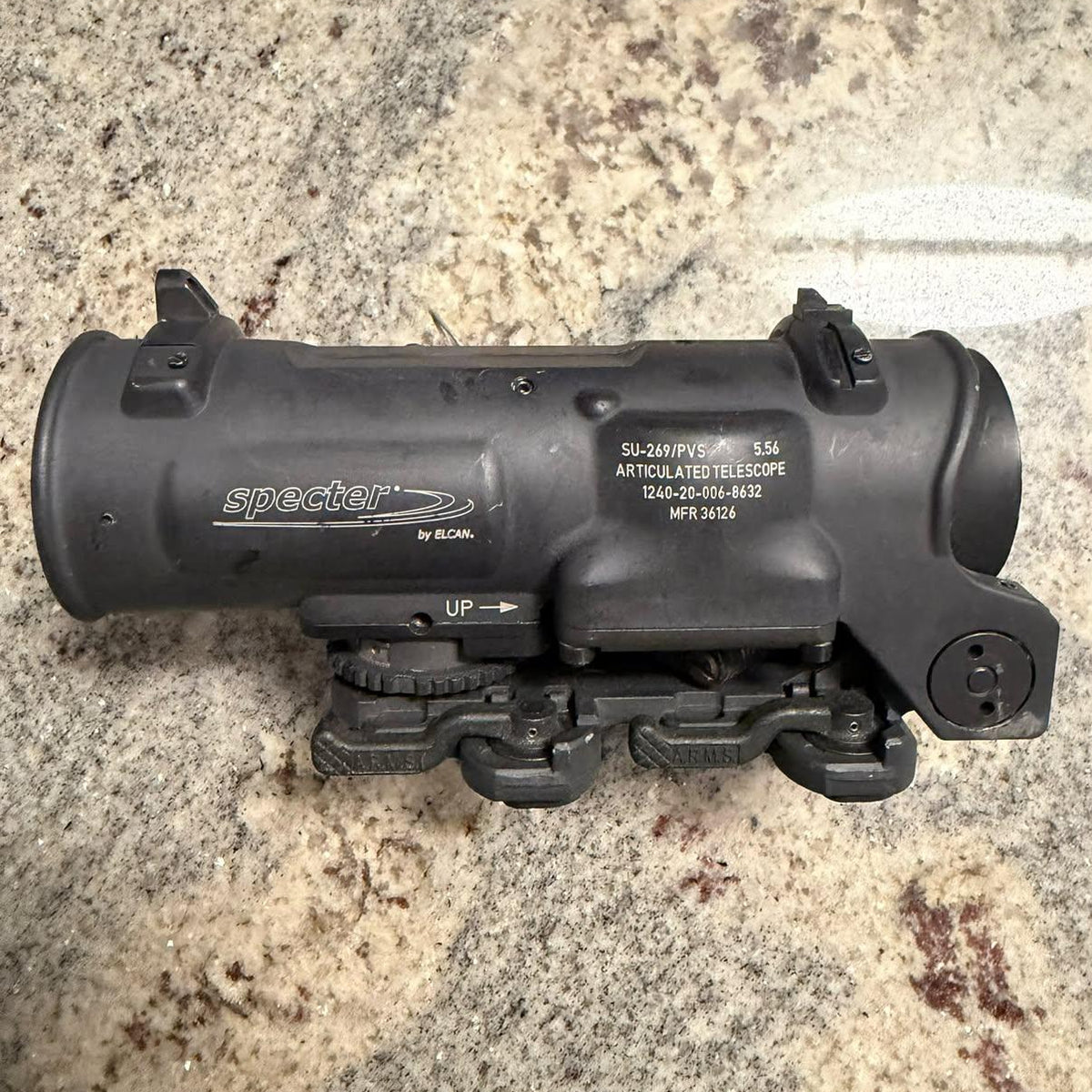 A black Specter AR-15 rifle scope photographed from above on a speckled beige and brown stone surface.
